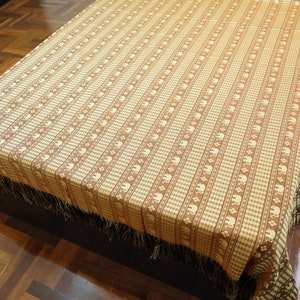 Puede incluir: Una manta tejida de color marrón y crema con un borde de flecos. La manta presenta un patrón repetitivo de pequeños elefantes blancos.