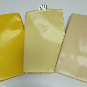 May include: Three folded fabric swatches in shades of yellow and beige. The swatches are labeled #9, #8, and #27. The fabric has a shiny, smooth texture, suggesting it may be satin or a similar material. The colors range from a bright yellow to a light beige.