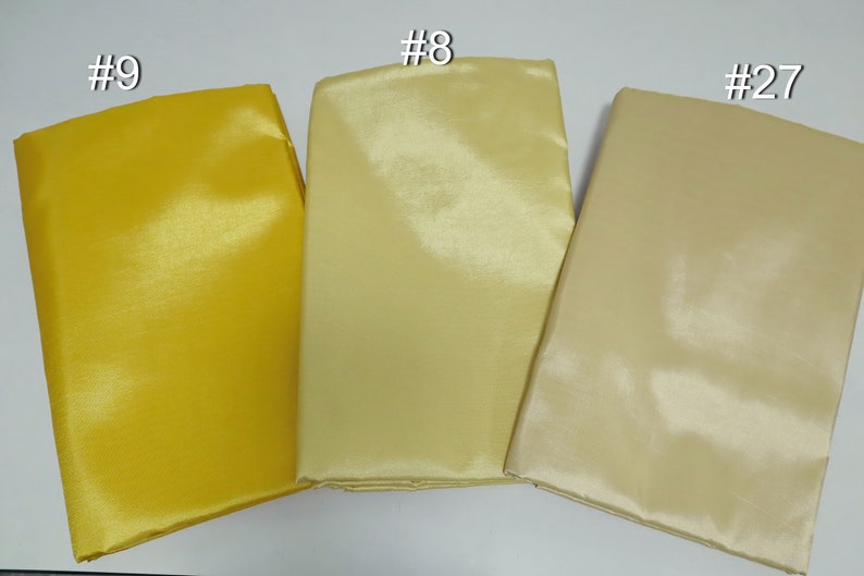 May include: Three folded fabric swatches in shades of yellow and beige. The swatches are labeled #9, #8, and #27. The fabric has a shiny, smooth texture, suggesting it may be satin or a similar material. The colors range from a bright yellow to a light beige.