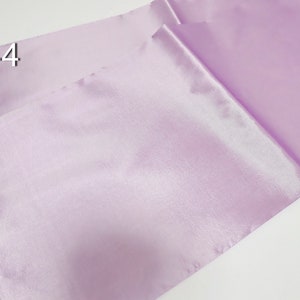 May include: A pair of lavender satin table runners with white lace trim. The runners are folded and angled, showcasing the fabric's sheen. The number "#44" is printed in white in the upper left corner. Ideal for weddings or special events.