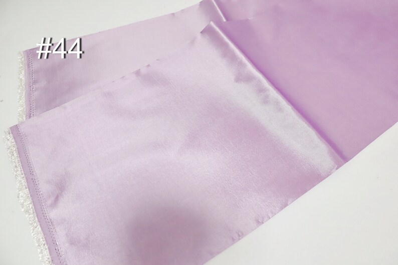 May include: A pair of lavender satin table runners with white lace trim. The runners are folded and angled, showcasing the fabric's sheen. The number "#44" is printed in white in the upper left corner. Ideal for weddings or special events.