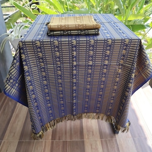 May include: A blue tablecloth with a gold elephant pattern and fringe. The tablecloth is folded over a stack of three gold and black patterned fabric squares.