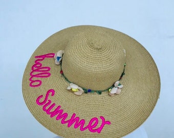 May include: A straw sun hat with a pink "Hello Summer" embroidery and a floral trim.