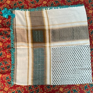 Puede incluir: Un pañuelo cuadrado estampado con base blanca, con detalles en marrón, turquesa y dorado. El pañuelo tiene un borde turquesa con flecos y un diseño geométrico. El pañuelo está doblado y expuesto sobre una superficie roja estampada.