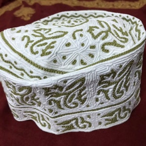 Puede incluir: Calota blanca con intrincados bordados verdes. El bordado presenta un patrón repetitivo de diseños florales.