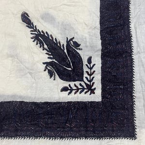 May include: A white fabric textile featuring a dark navy blue embroidered border and a matching dark navy blue embroidered floral motif in the corner. The embroidery consists of dense, textured stitching that creates a visible pattern on the surface.
