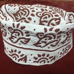 Puede incluir: Una gorra de algodón blanca con un diseño bordado marrón. La gorra tiene una parte superior redondeada y una visera plana.