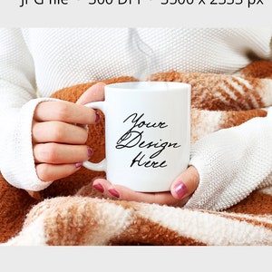 May include: A white mug held by a person, with the text "Your Design Here" in black script. The mug is set against a cozy backdrop of a white sweater and a brown and white patterned blanket. The image also includes text that says "MUG MOCKUP".