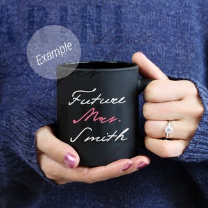 May include: A black ceramic mug with the words "Future Mrs. Smith" in white and pink script. The mug is held by a person wearing a blue sweater. A diamond ring is visible on the person's finger.