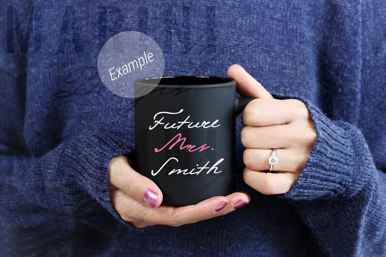May include: A black ceramic mug with the words "Future Mrs. Smith" in white and pink script. The mug is held by a person wearing a blue sweater. A diamond ring is visible on the person's finger.