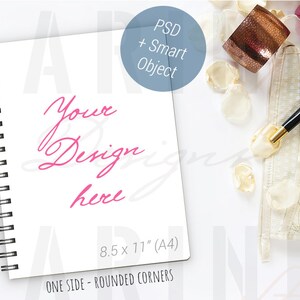 K&ouml;nnte beinhalten: Wei&szlig;es Spiralnotizbuch mit dem rosa Schriftzug "Your Design here". Das Notizbuch ist 21,6 x 27,9 cm (A4) gro&szlig; und hat abgerundete Ecken. Ein schwarz-goldener Stift, eine dekorative Manschette und Bl&uuml;tenbl&auml;tter sind ebenfalls vorhanden.