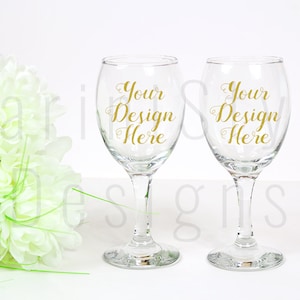 Puede incluir: Dos copas de vino transparentes con el texto "Your Design Here" en escritura dorada. Las copas están sobre un fondo blanco con un ramo de flores blancas a la izquierda. Ideal para diseños personalizados.