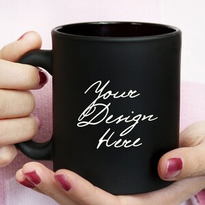 May include: A black mug with the text "Your Design Here" in a white script font. The mug is held by hands with red nail polish. The words "BLACK MUG MOCKUP" are at the top.
