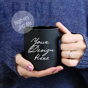 May include: A black mug with the text "Your Design Here" in white script. The mug is held by a person wearing a blue sweater. The image includes a ring with a diamond.