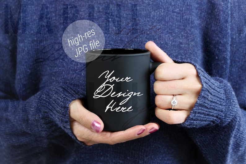 May include: A black mug with the text "Your Design Here" in white script. The mug is held by a person wearing a blue sweater. The image includes a ring with a diamond.