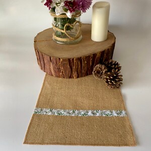 Op de afbeelding: Een jute tafelkleed met een wit lint met een groen bladmotief. De loper wordt getoond op een houten boomstamschijf met een glazen vaas met paarse en witte bloemen en een witte kaars.