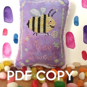 Puede incluir: Una almohada de punto de cruz de color morado con un diseño de abeja y el texto "Bee Sweet". La almohada está rodeada de gominolas.