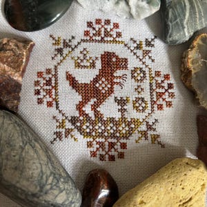 May include: A cross-stitch design featuring a brown dinosaur within a circular frame, surrounded by decorative floral elements in shades of brown, yellow, and orange. The design is set against a white fabric background, with various stones around the edges.