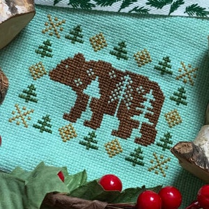 May include: A cross-stitch pattern of a brown bear on a light blue background. The bear is surrounded by a border of green trees, gold stars, and white snowflakes.