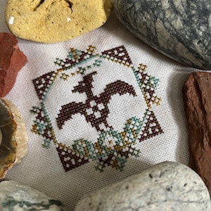 May include: Cross-stitch embroidery featuring a stylized dragon in shades of brown, surrounded by a square border and floral accents in teal and gold. The design is on a cream-colored fabric.