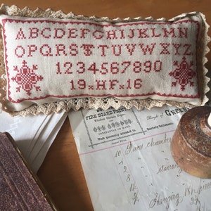 May include: A cream-coloured cross-stitch sampler cushion with red lettering and numbers, edged with beige lace. The sampler displays the alphabet and numbers. A vintage ink bottle and aged documents are also visible in the image.