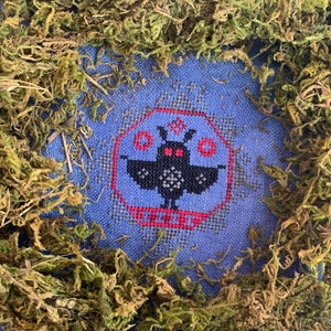 May include: A blue fabric with a cross-stitch design of a black bat with red eyes. The bat is surrounded by a red and white border. The fabric is framed by a border of dried moss.
