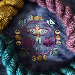 May include: A cross-stitch pattern featuring a butterfly with a yellow body and green wings, surrounded by a floral design. The pattern is stitched on a dark blue fabric. Three skeins of yarn in teal, purple, and mustard yellow are arranged around the pattern.
