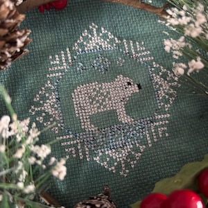 May include: A teal blue cross-stitch embroidery of a white polar bear standing on a blue and white snowflake design. The embroidery is framed by a brown twig wreath with red berries and white pine cones.
