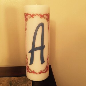 May include: White pillar candle with a red and white decorative border and the letter "A" in blue in the center.