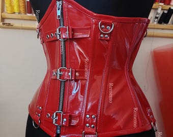 May include: A glossy red corset featuring a vertical front zipper closure, three horizontal buckled straps, and multiple silver-toned metal D-rings attached to the front and sides. The garment has a structured, shiny finish with visible stitching.