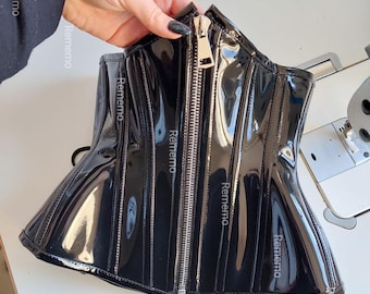 May include: A black corset with a glossy, patent-like finish featuring a prominent silver-toned metal zipper running down the center front. The garment is constructed with vertical panels and includes black lacing at the bottom edge.