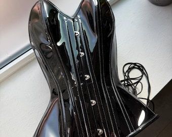 May include: A black corset made of a glossy, patent-like material. The garment features a structured, paneled design with a sweetheart neckline and a vertical row of silver-toned metal busk closures down the center front. Long black laces are attached.