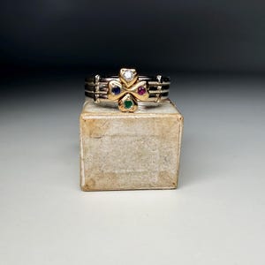 Puede incluir: Un anillo de varias bandas con una pieza central en forma de trébol hecha de oro amarillo. El trébol está engastado con una piedra blanca transparente en la parte superior, un zafiro azul a la izquierda, un rubí rojo a la derecha y una esmeralda verde en la parte inferior.