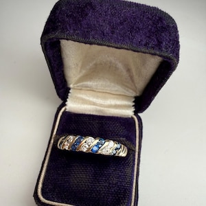 May include: A gold ring with alternating blue and clear stones, presented in a dark purple velvet ring box with a cream-colored satin interior. The ring's design features a repeating pattern of gemstones.