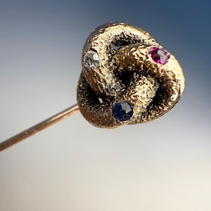 May include: A gold-toned tie pin in a knot design, adorned with a clear diamond, a ruby, and a sapphire. The pin has a textured surface and a slender, gold-colored stem.