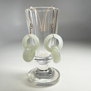May include: A pair of jade earrings with gold-colored hooks. Each earring features a large, pale green circular element connected to a smaller, similar element. The earrings are displayed in a clear glass.