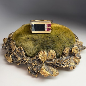 May include: A gold ring with a rectangular black onyx center and ruby accents, resting on a green velvet cushion with a gold-toned, ornate base. The ring's design is geometric and bold.