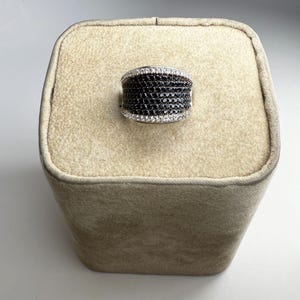 May include: A wide, silver-toned ring with a band of small, black stones. The top and bottom edges are lined with a row of smaller, white stones. The ring is displayed on a beige, rectangular stand.
