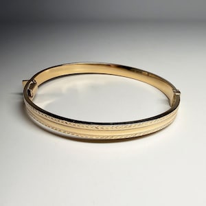 May include: A gold-toned hinged bangle bracelet featuring a polished centre band flanked by two textured, cross-hatched decorative borders. The bracelet includes a secure clasp mechanism on one side and a smooth, rounded interior profile.