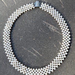 Könnte beinhalten: Eine weiße Perlenkette mit einem mehrsträngigen Design. Die Halskette besteht aus zahlreichen kleinen, runden Perlen, die ein strukturiertes, geschichtetes Aussehen erzeugen. Der Verschluss ist silberfarben. Die Halskette wird auf einer dunkelgrauen Oberfläche präsentiert.