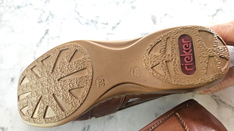 May include: Brown leather sandal with a textured sole. The sole features a geometric pattern and the brand name "riecker" in a red oval. The shoe size is 39.