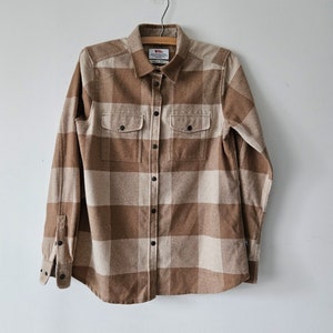 May include: A brown and white plaid flannel shirt with a button-up front and two chest pockets. The shirt is hanging on a wooden hanger.