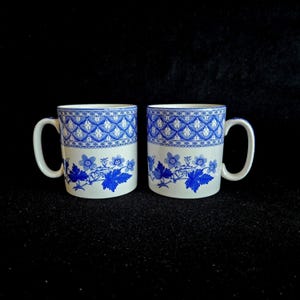 May include: Two white ceramic mugs with blue floral and geometric patterns. The mugs have a handle and a blue and white design around the top. The bottom of the mugs feature blue leaves and flowers.