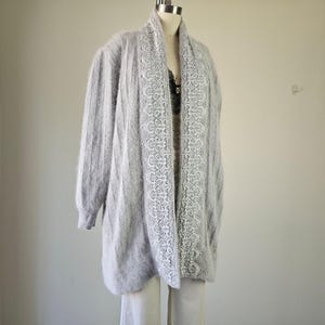 Può includere: Un cardigan grigio chiaro a maniche lunghe con un bordo in pizzo bianco sul davanti. Il cardigan è esposto su un manichino. Il capo ha una consistenza morbida e sfocata e una vestibilità ampia.