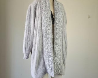 May include: A light gray, long-sleeved cardigan with a white lace trim down the front. The cardigan is displayed on a mannequin. The garment has a soft, fuzzy texture and a relaxed fit, suitable for layering.