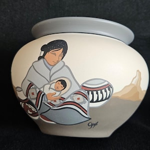 May include: A hand-painted ceramic pot with a grey rim and a cream-coloured body. The pot features a Native American woman holding a baby, with a stylised landscape and a patterned pot in the background. The artist's signature is visible.