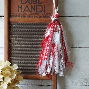 May include: A decorative tassel with red and white ribbons and a key charm hangs in front of a vintage wooden washboard. A mason jar filled with dried flowers and a wooden sign that reads "Hudson River Craftings" are also in the image.