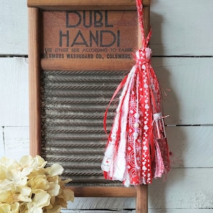 May include: A white washboard with red and white fabric tassel hanging from it. The washboard has the text "DUBL HANDI" and "COLUMBUS WASHBOARD CO., COLUMBUS" printed on it. The washboard is sitting on a wooden surface. There is a white jar with a dried flower arrangement in front of the washboard. The jar has the word "DEAL" printed on it. There is a wooden sign with the text "Hudson River Craftings" in front of the jar.