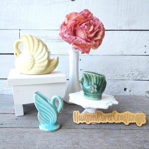 May include: A still life arrangement featuring three ceramic swan-shaped vases in yellow, teal, and green. A large pink flower sits in a white vase. The vases are on a white wooden surface with a wooden sign that reads "Hudson River Craftings."