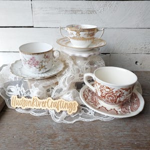 May include: Several antique teacups and saucers are arranged on a lace cloth. One cup features a floral design, while another has a brown transferware pattern. A wooden sign with the text "HudsonRiverCraftings" is visible.